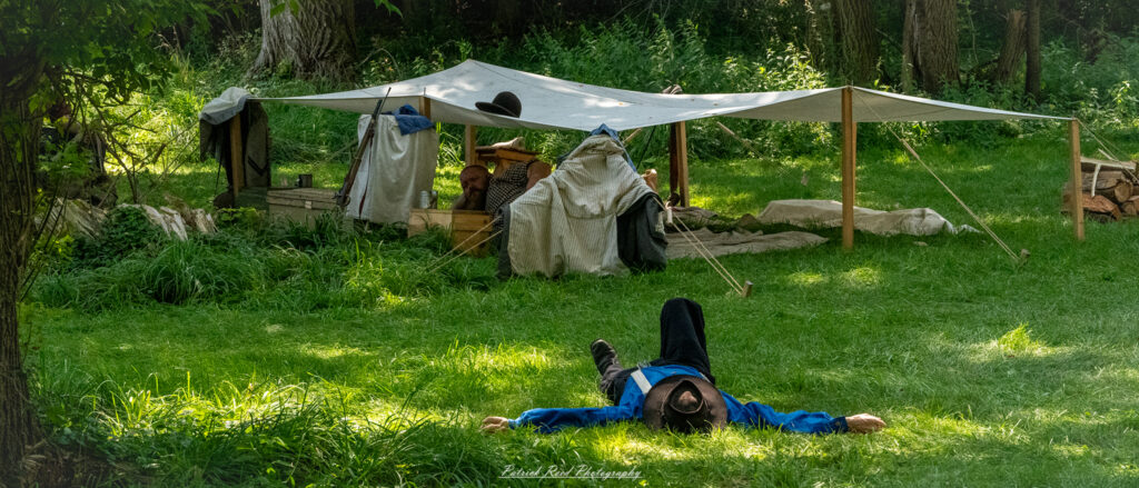 A soldier napping on the grass, dressed in a military uniform, with his rifle resting nearby. The scene is peaceful, with sunlight filtering through the trees, creating a serene atmosphere as he takes a moment of rest amidst the demands of duty.